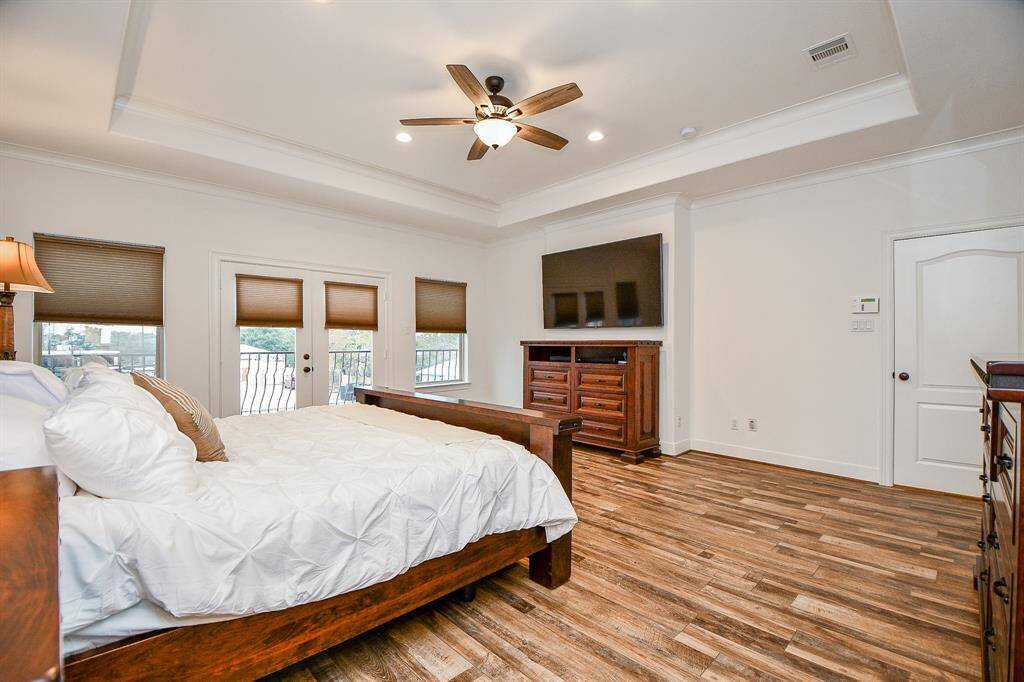 Spacious bedroom with wood flooring, large bed, ceiling fan, and a mounted TV. Features ample natural light from multiple windows and French doors leading to a balcony.