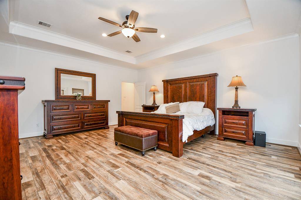 This spacious bedroom features elegant hardwood floors, a high ceiling with a fan, and ample natural light. The room is furnished with a large wooden bed, matching nightstands, and a dresser with a mirror, creating a warm and inviting atmosphere.