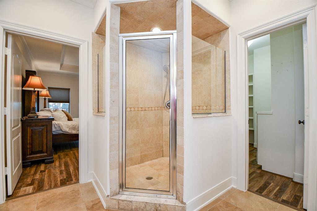 The photo shows a modern bathroom setup with a central glass-enclosed shower featuring beige tiles. To the left is a view into a cozy bedroom with wooden flooring, and to the right is an entryway to a walk-in closet. The space is well-lit and has a warm, inviting feel.