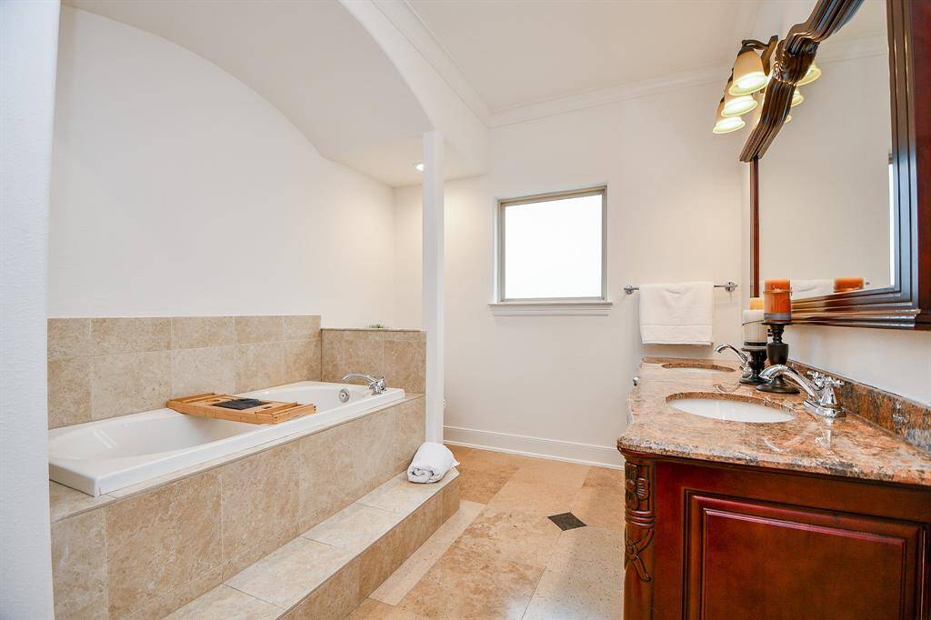 This spacious bathroom features a luxurious soaking tub with a tile surround, a dual-sink vanity with a granite countertop, and elegant lighting. It also has a window for natural light and a warm, inviting color palette.