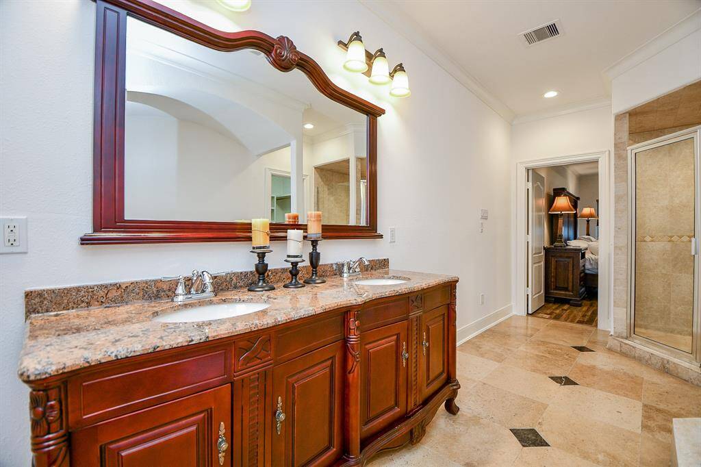 This bathroom features a double vanity with a granite countertop, elegant wood cabinetry, and a large framed mirror. It includes a spacious shower and connects to a bedroom with a warm, inviting atmosphere.