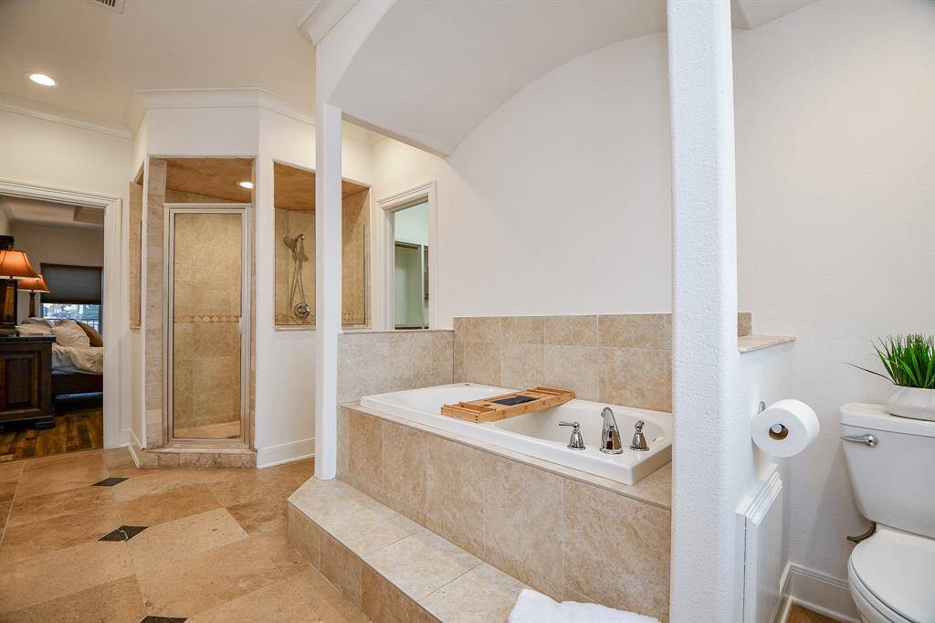 This spacious bathroom features a tiled soaking tub with modern fixtures, a separate glass-enclosed shower, and a clean, natural aesthetic. The adjoining bedroom visible adds convenience and accessibility.
