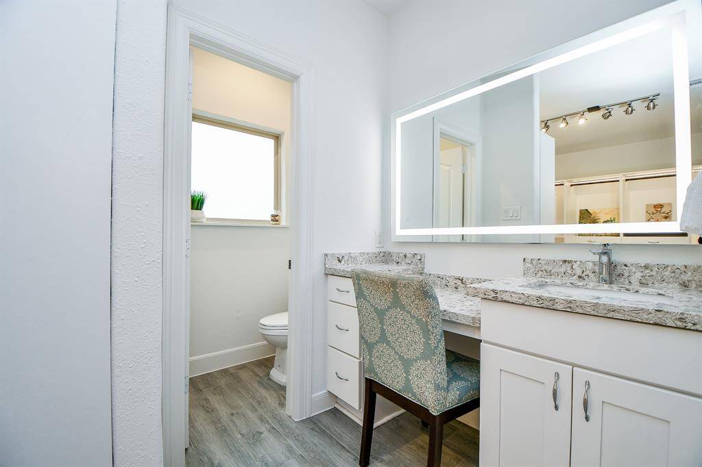 This modern bathroom features a sleek vanity with a large, illuminated mirror, granite countertops, and ample cabinet space. The space includes a separate toilet area with a window for natural light, and stylish wood-like flooring.