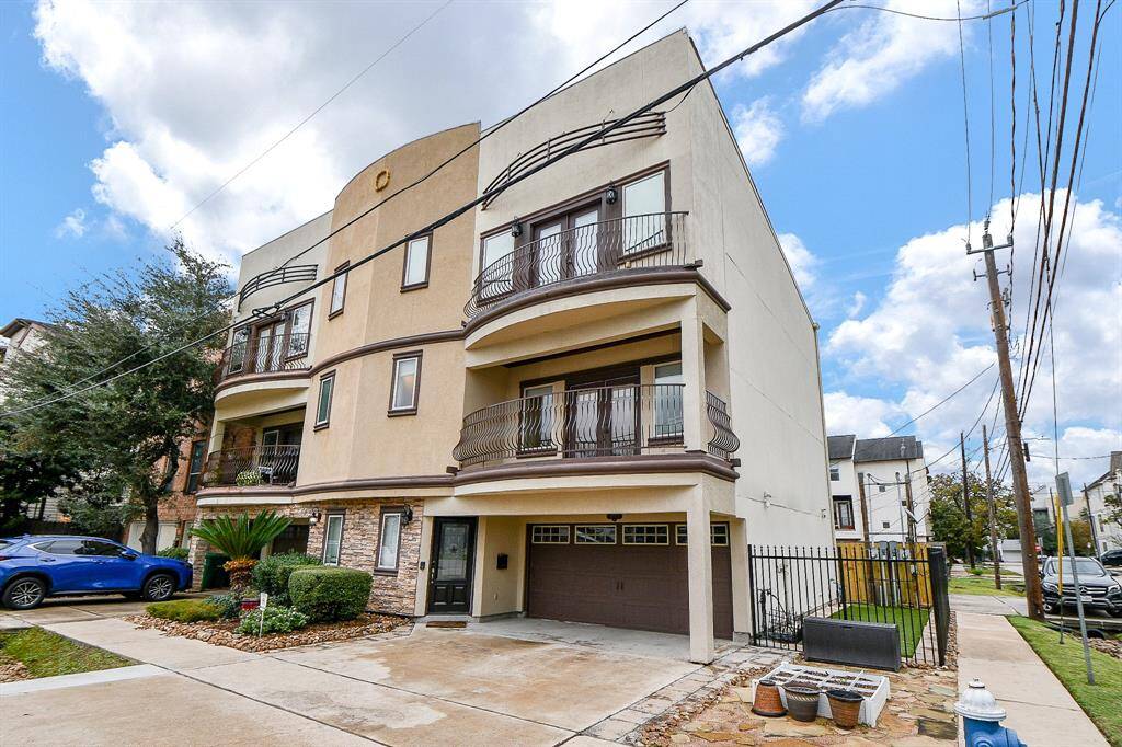 This modern three-story townhouse features a stucco exterior with stone accents, two balconies, and a spacious two-car garage. The property is complemented by a small landscaped area and a driveway, situated in a residential neighborhood with nearby sidewalks and street access.