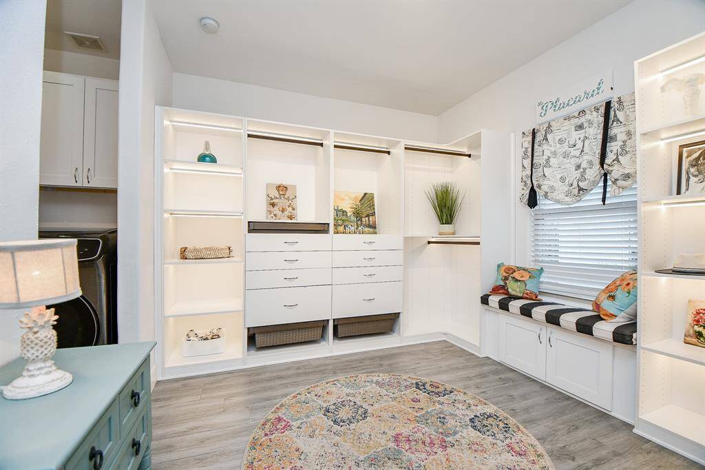This photo shows a bright, organized laundry room with built-in cabinets and shelving. It features a window seat with colorful cushions, a patterned rug, and ample storage space, creating a functional and inviting area.