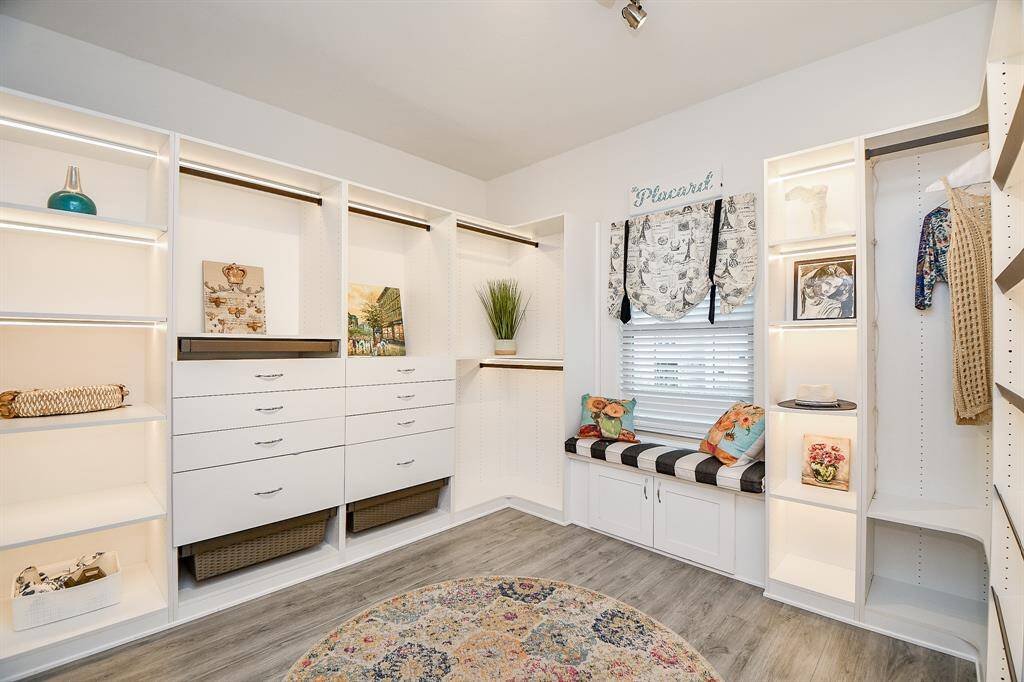 This photo showcases a spacious walk-in closet with ample storage including open shelves, hanging space, and drawers. It features a cozy window seat with cushions, perfect for relaxing. The room has bright lighting and a stylish, neutral palette, ideal for organizing and enjoying your wardrobe.