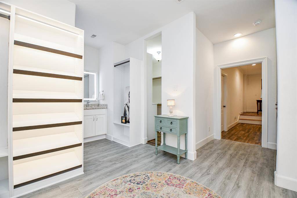 This bright, spacious room features laminate wood flooring and ample built-in shelving, perfect for storage or display. There's a small, stylish table with a lamp, and the area leads into a hallway with more space visible in the background. Ideal for a home office or study.