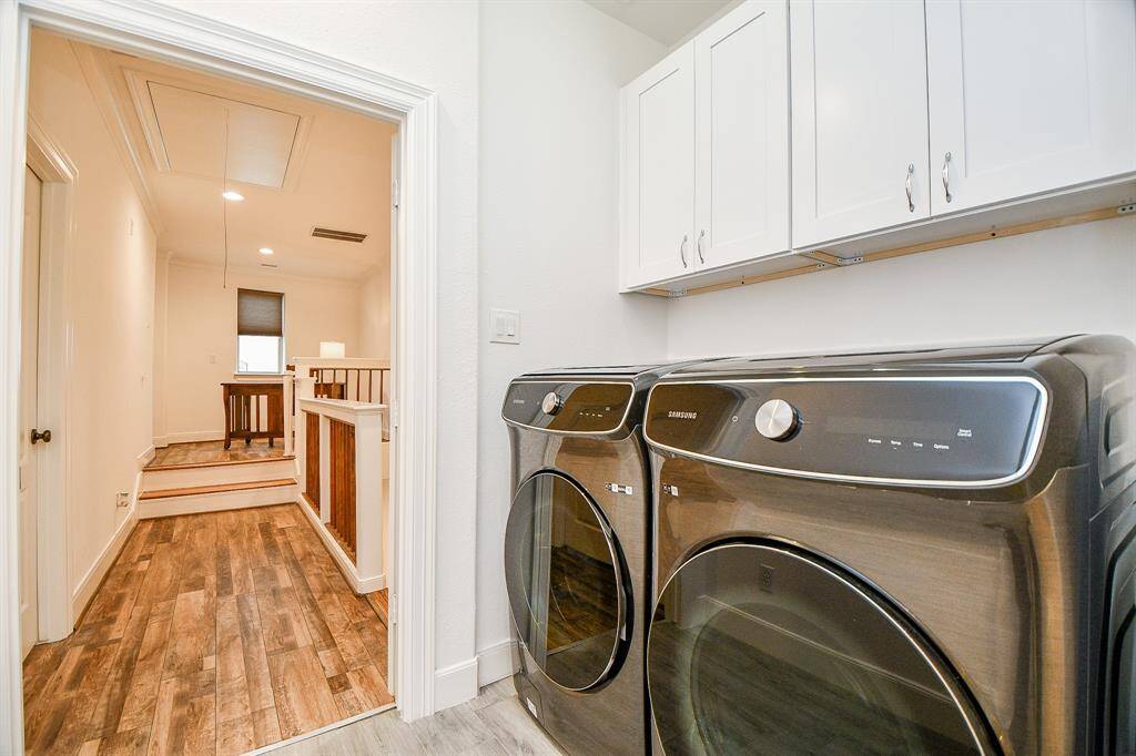 This photo shows a bright laundry area with modern Samsung washer and dryer units. It has white cabinetry above for storage and wood-look flooring. The space opens to a hallway with stairs leading up to a cozy area with natural light.