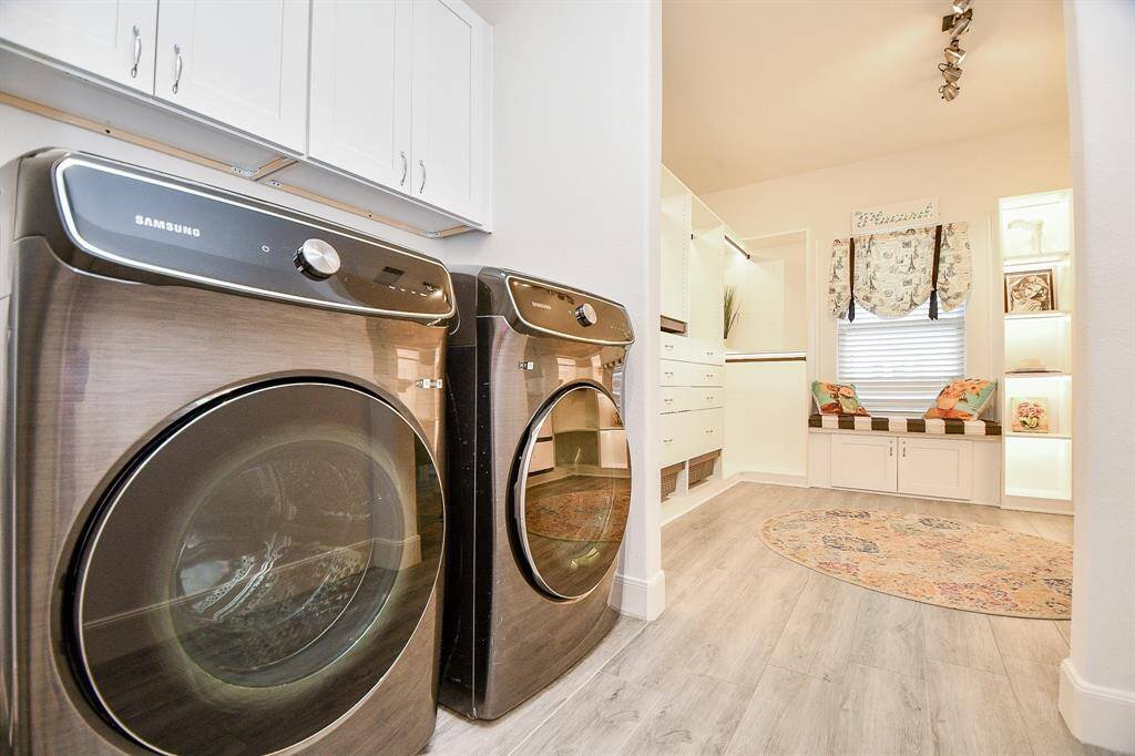 This photo showcases a bright, modern laundry room with sleek Samsung washer and dryer units. It features ample cabinet storage, a cozy window seat with cushions, and open shelves for display. The space is well-lit, with light wood flooring and a decorative rug adding warmth.