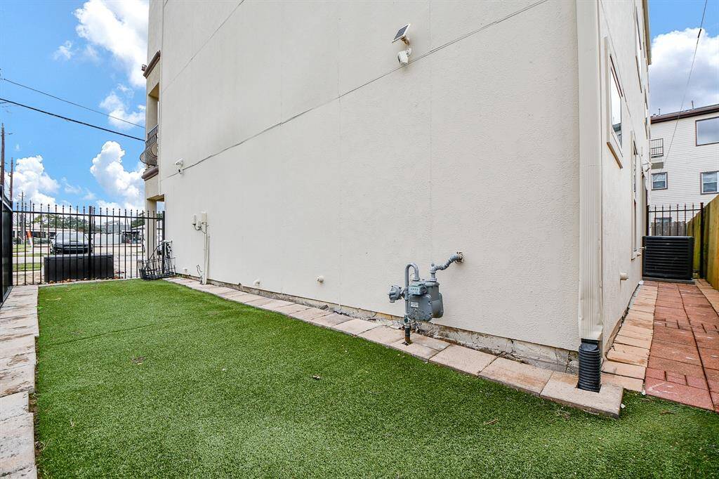 This photo shows a side yard with artificial turf, bordered by a light-colored building wall. There's a paved walkway and a black metal fence providing enclosure. The area is simple and low-maintenance, suitable for outdoor activities or pets.