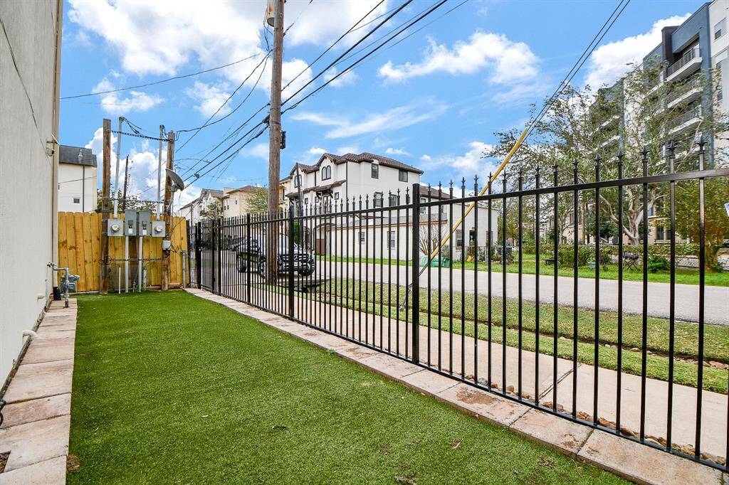 This photo shows a narrow side yard with artificial turf, bordered by a black metal fence. It includes utility meters and is adjacent to a street with nearby residential buildings.