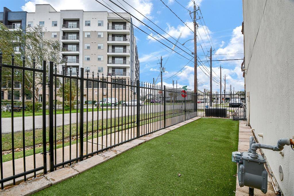 This photo shows a fenced side yard with artificial grass, located next to a modern apartment building. The area is bordered by a sidewalk and a street, with some visible utility installations and power lines overhead.