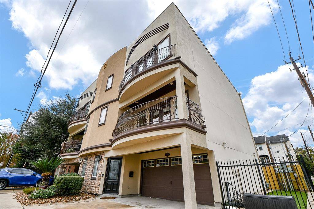 Modern three-story townhouse with a unique curved facade and two balconies. Features a two-car garage and gated entry, with a small landscaped front area. Located in a neighborhood with nearby utility poles and other multi-story homes.