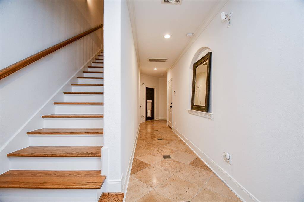 This photo shows a bright and inviting hallway featuring tiled flooring, a staircase with wooden steps, and a wall-mounted mirror. The space is well-lit with recessed lighting.
