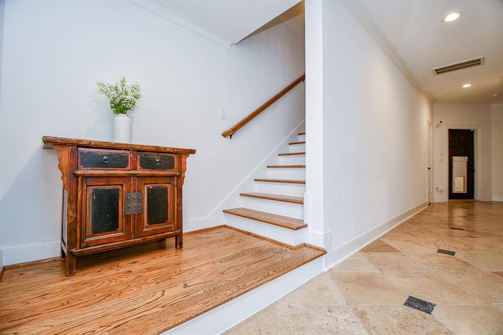 This inviting entryway features a wooden staircase with a classic handrail, a tiled floor, and a small vintage cabinet with decorative accents. The space is bright and open, offering a welcoming atmosphere.
