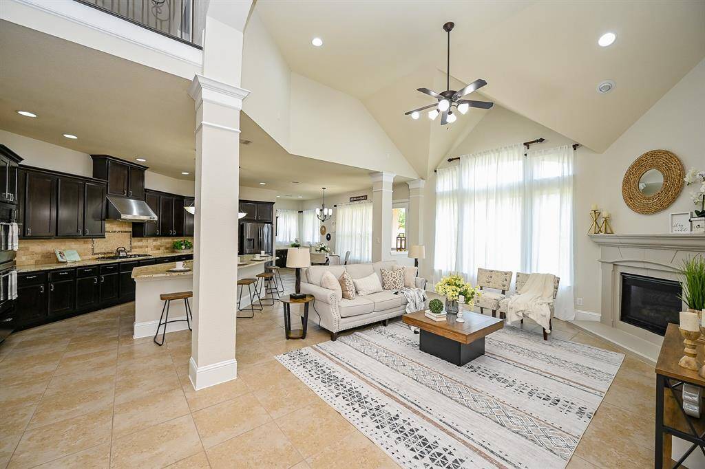 A bright, open-concept living area with high ceilings, large windows, and elegant tile flooring. The space includes a cozy living room with a fireplace, modern kitchen with dark cabinetry and an island, and stylish decor throughout. Perfect for comfortable living and entertaining.