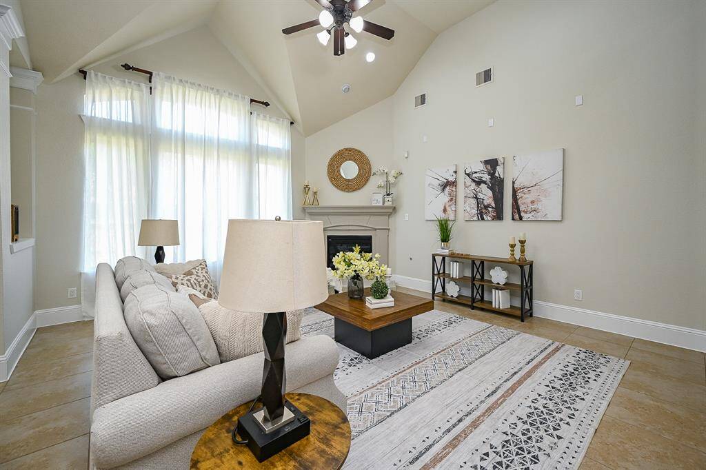 This spacious living room features high vaulted ceilings, a cozy fireplace, and large windows for ample natural light. The neutral tones and elegant decor create a warm, inviting atmosphere perfect for relaxation or entertaining.