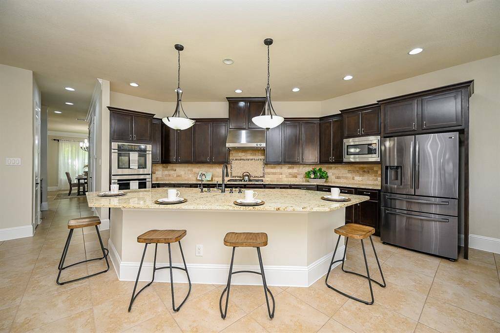 This modern kitchen features dark wood cabinetry, stainless steel appliances, and a large island with granite countertops. Pendant lighting and a tile backsplash add elegance, while an open layout connects to the dining area.