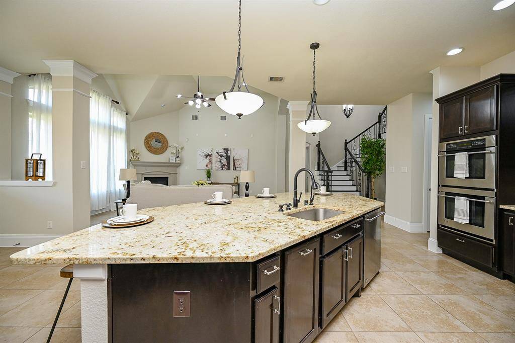This spacious kitchen features a large granite island with a sink and seating, elegant pendant lighting, dark wood cabinetry, and stainless steel appliances. It opens to a bright living area with high ceilings, a fireplace, and access to a staircase, creating an inviting and functional space.