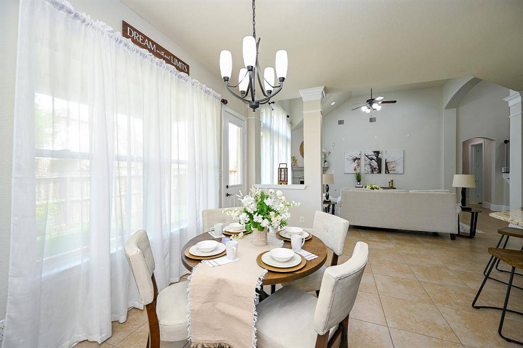The breakfast space with soft natural light filtering through sheer curtains. The room extends into a comfortable living area with neutral tones, tiled flooring, and elegant lighting fixtures.