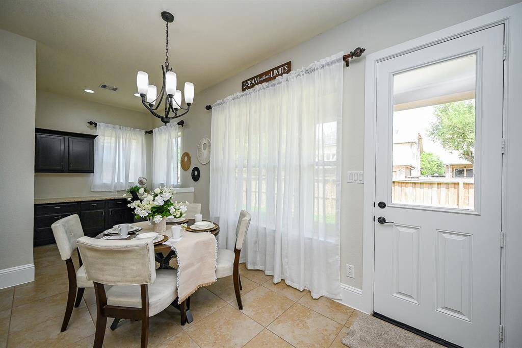 Bright breakfast area with elegant chandelier, neutral tones, and tiled floors. Features large windows with sheer curtains, and a door leading to the backyard. Ideal for intimate gatherings.