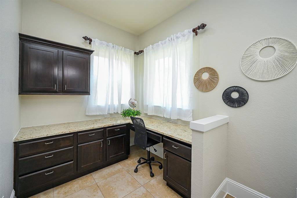 A small, bright home office with a corner desk and dark wood cabinetry. It has tiled flooring, two windows with sheer curtains, for a stylish touch.