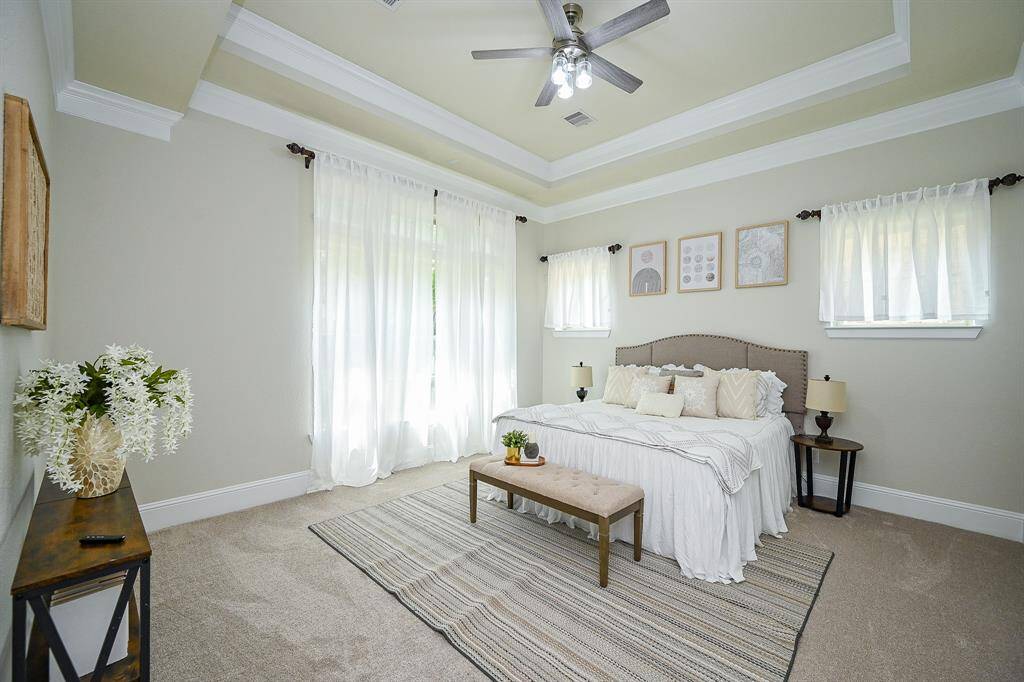 Bright and airy primary bedroom with a neutral palette, featuring a tray ceiling, ceiling fan, and plush carpet. Large windows with sheer curtains allow natural light, complemented by tasteful wall art and cozy furnishings.