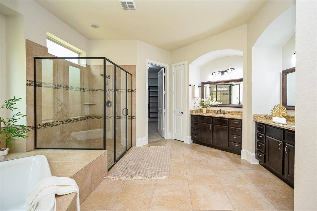 The spacious primary bathroom features a large glass-enclosed shower, a separate bathtub, dual sinks with granite countertops, and elegant dark wood cabinetry. Natural light streams in, enhancing the neutral tones and creating a serene, spa-like atmosphere.