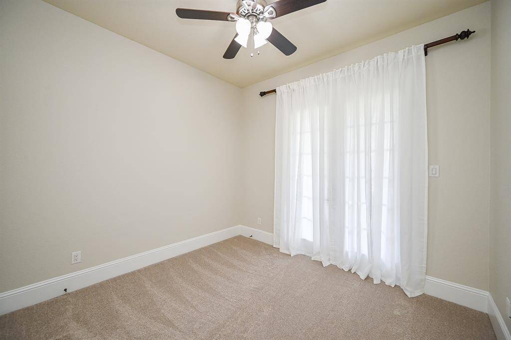 This second bedroom down features neutral walls, carpeted flooring, a ceiling fan, and large windows with sheer curtains, offering ample natural light.