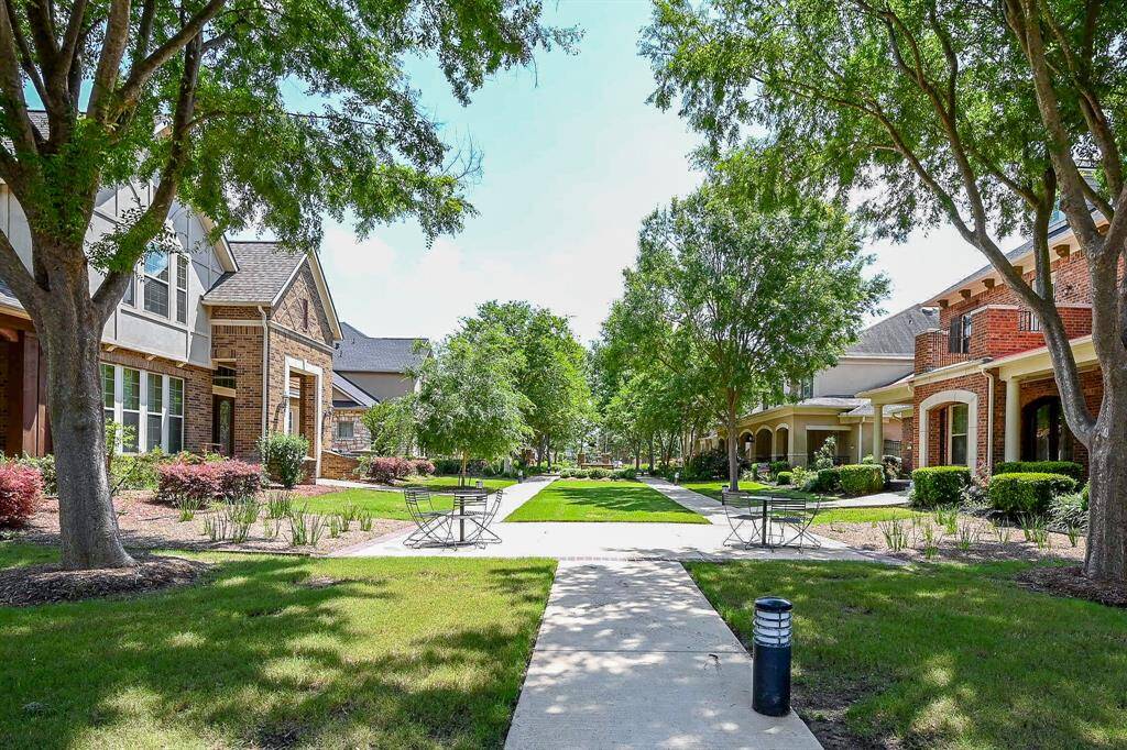 A charming neighborhood with well-maintained homes, lush landscaping, and mature trees lining a central walkway. The inviting outdoor seating and serene atmosphere create a welcoming community environment.