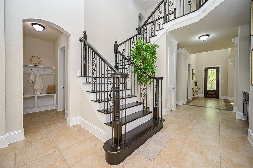 A bright, spacious foyer with elegant tile flooring and a grand staircase featuring wrought iron railings. The entrance leads to a well-lit hallway with high ceilings, and there's a cozy nook with storage. A touch of greenery adds warmth to the space.