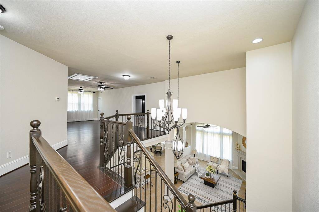 A spacious and elegant two-story home interior. It features a view from the staircase landing with an ornate railing, overlooking a bright living room with high ceilings and large windows. A modern chandelier adds sophistication, and the hardwood floors contribute to the warm ambiance.