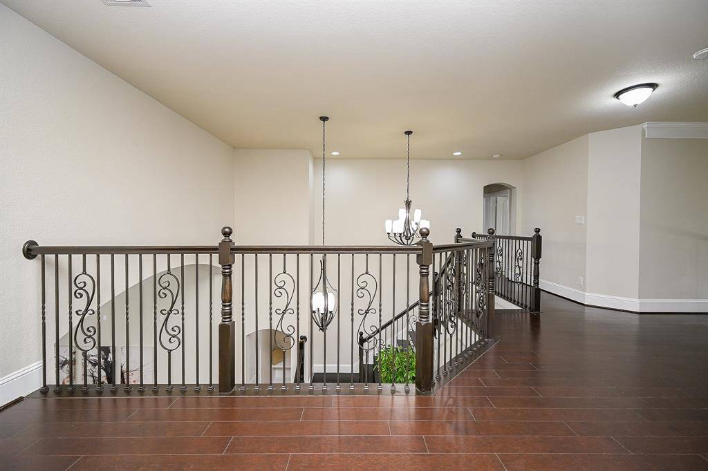 A spacious, elegant upstairs landing with dark hardwood floors and decorative wrought iron railings. The area is well-lit by a stylish chandelier and recessed lighting, offering a sophisticated and open feel.