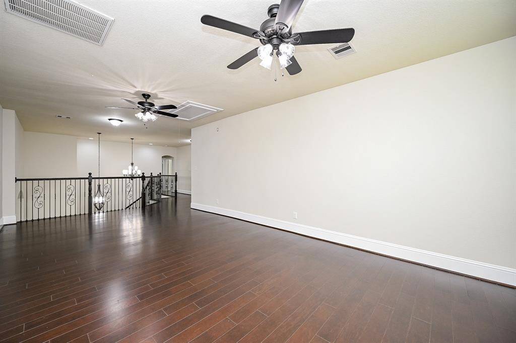Spacious, bright game room with dark hardwood floors and neutral walls, featuring ceiling fans and elegant iron railing overlooking a lower level.