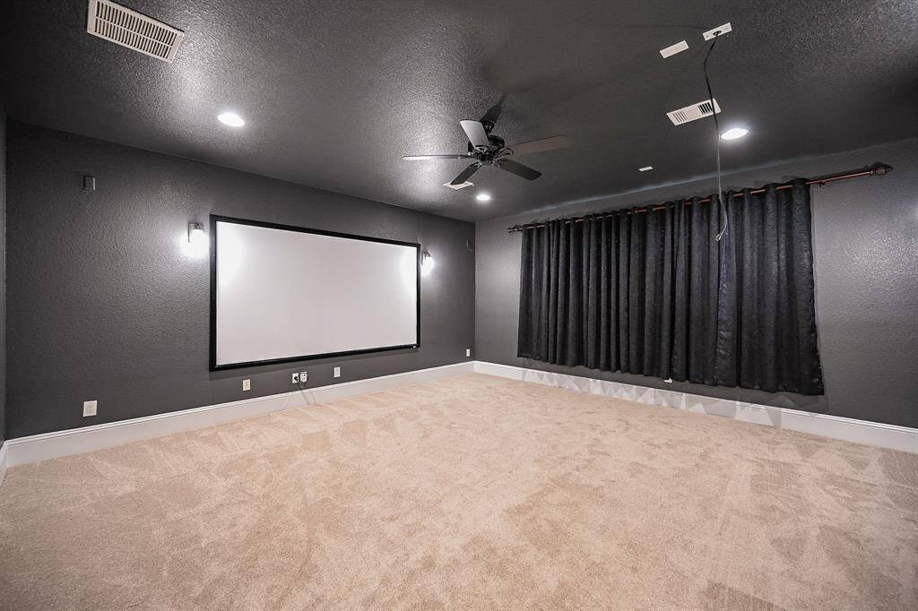 A spacious home theater room with dark walls, a ceiling fan, and recessed lighting. It features a large screen, carpeted flooring, and blackout curtains, perfect for an immersive viewing experience.