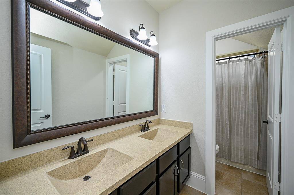 This bathroom features a double sink vanity with a large framed mirror, elegant faucets, and ample cabinetry. It includes a separate area with a shower-tub combination and neutral tile flooring, providing a modern and functional space.