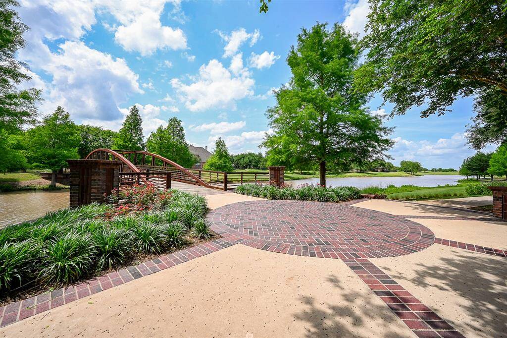 This photo showcases a serene outdoor space with a red brick path leading to a charming arched bridge over a small waterway. Lush greenery and mature trees surround the area, with a picturesque lake visible in the background, offering a peaceful and inviting setting.