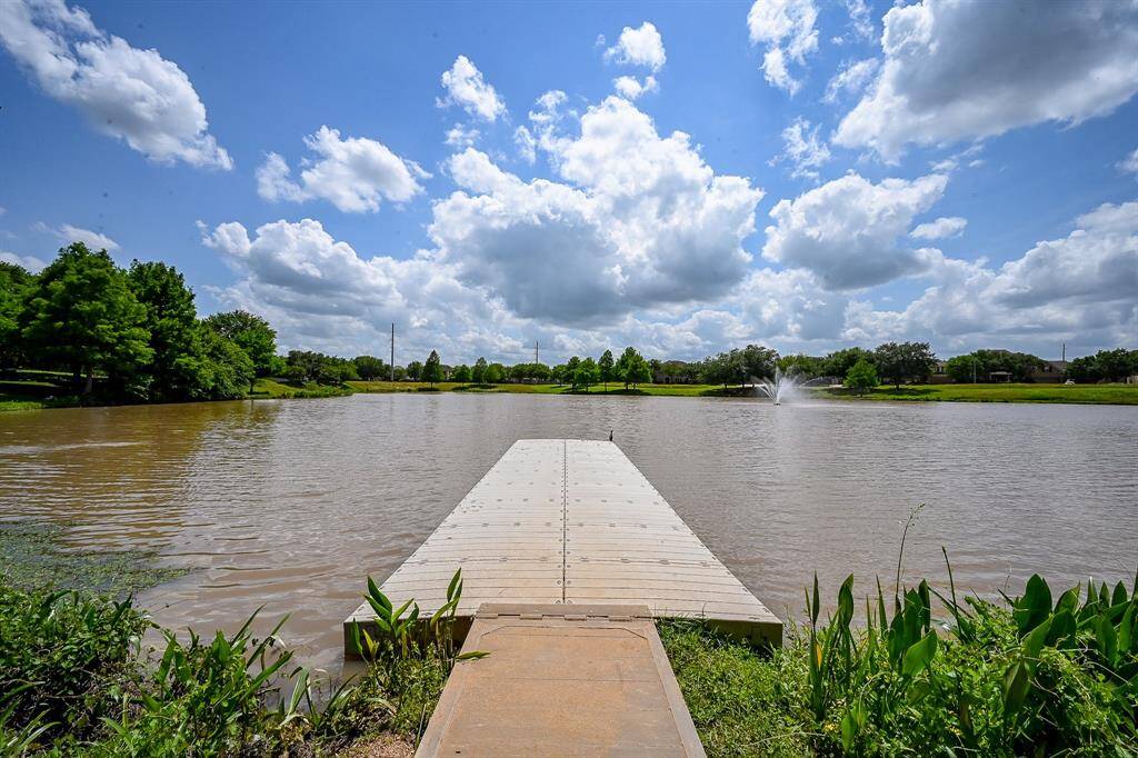 This community photo showcases a serene lakeside view with a wooden dock extending into the water, surrounded by lush greenery. The sky is bright with scattered clouds, and there's a fountain in the distance, creating a peaceful and inviting atmosphere.