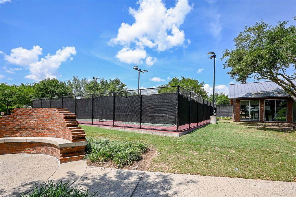 This photo showcases a well-maintained outdoor tennis court surrounded by greenery, with a nearby brick structure and a building, ideal for recreational activities in a community setting.