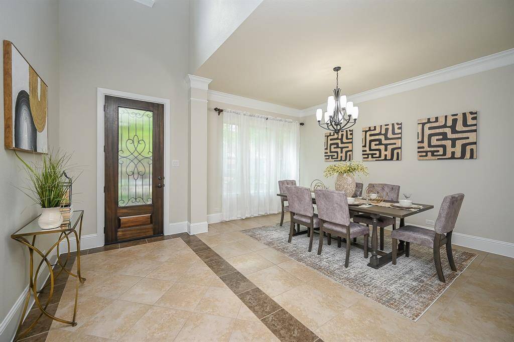 A bright, elegant dining area with a stylish chandelier and modern decor. The space features a large dining table with comfortable seating, tiled flooring, and a decorative entry door with glass details. Natural light streams in through sheer curtains, enhancing the room's inviting atmosphere.