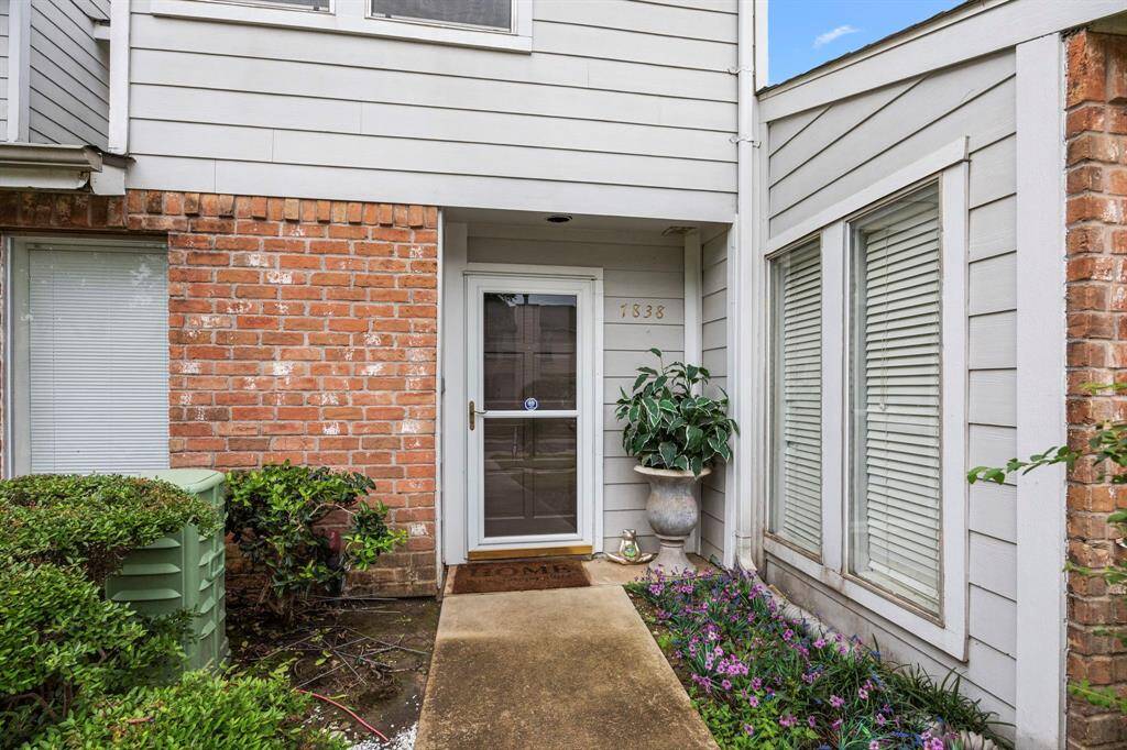 This photo shows the entrance of a home with a brick and siding exterior. The doorway is accompanied by a glass storm door, potted plants, and a welcome mat. The area is landscaped with shrubs and flowers.