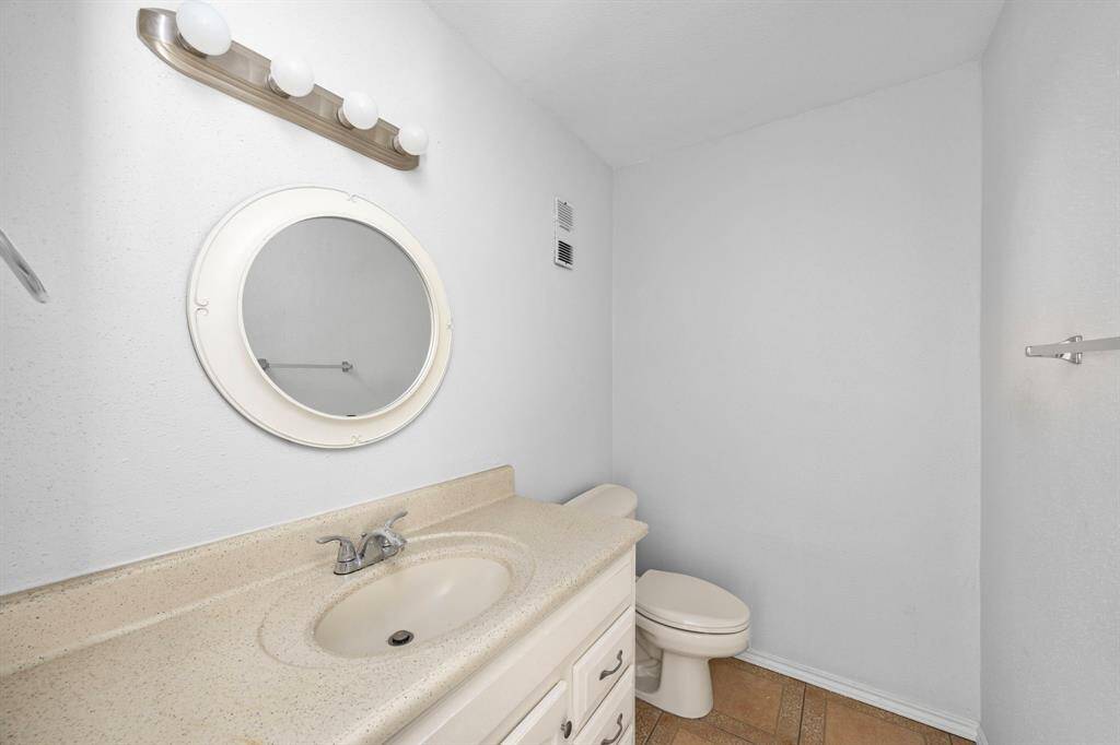 This bathroom features a simple design with a round mirror, a light fixture above, a single sink with a beige countertop, and a toilet. The space is compact with light-colored walls and tile flooring.