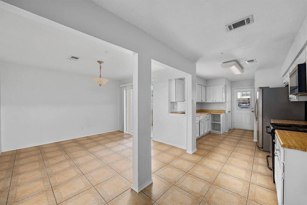 This photo showcases a bright open-concept kitchen and dining area. The kitchen features white cabinets, modern stainless steel appliances, and wooden countertops. The dining space is defined by a hanging light fixture and large sliding doors, offering plenty of natural light. The tiled floors and neutral walls create a clean, spacious feel.