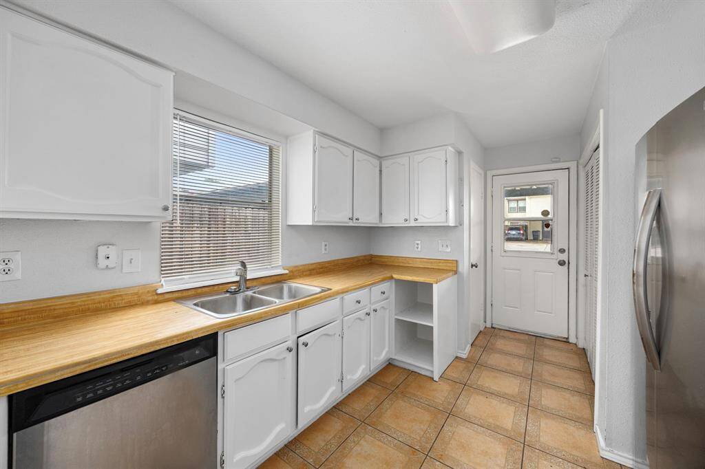 This kitchen features white cabinetry and light wood countertops, complemented by a double sink and stainless steel appliances. Natural light streams in through a large window, highlighting the tile flooring. A door at the end leads outside, adding convenience.