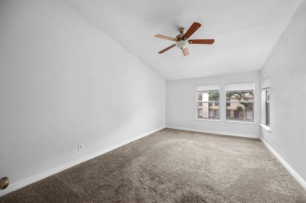 This spacious room features neutral carpeting, a high ceiling with a ceiling fan, and large windows that provide ample natural light and a view of the outdoors.