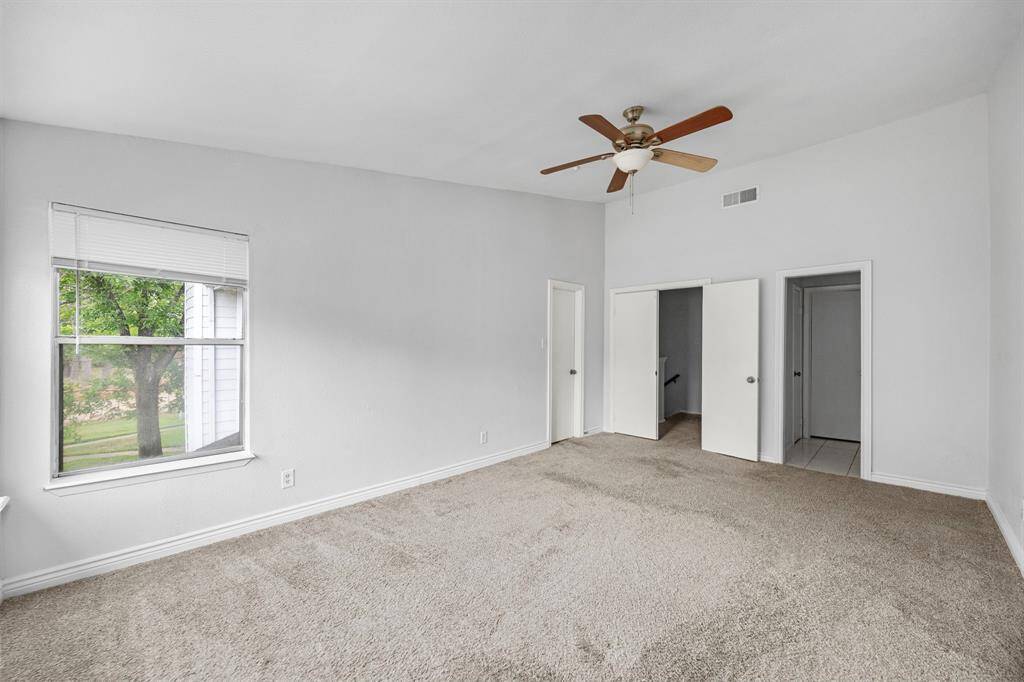 This spacious bedroom features neutral carpeting, a ceiling fan, and ample natural light from a large window. It includes two closets and a doorway leading to additional rooms, offering versatile storage and layout options.