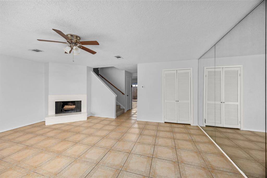 This spacious living area features tiled flooring, a cozy fireplace, a ceiling fan, and mirrored closet doors. It also includes a staircase leading to another level, offering a bright and open layout.