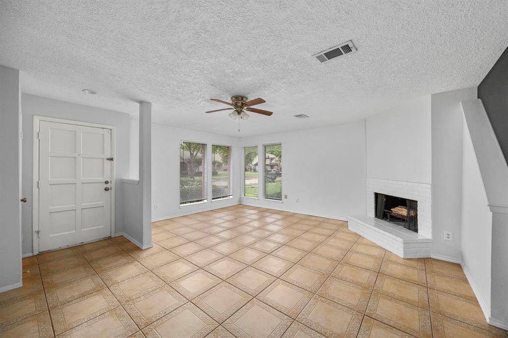 Bright living area with large windows, tile flooring, and a cozy corner fireplace. The room features a ceiling fan and neutral walls, offering a welcoming and versatile space.