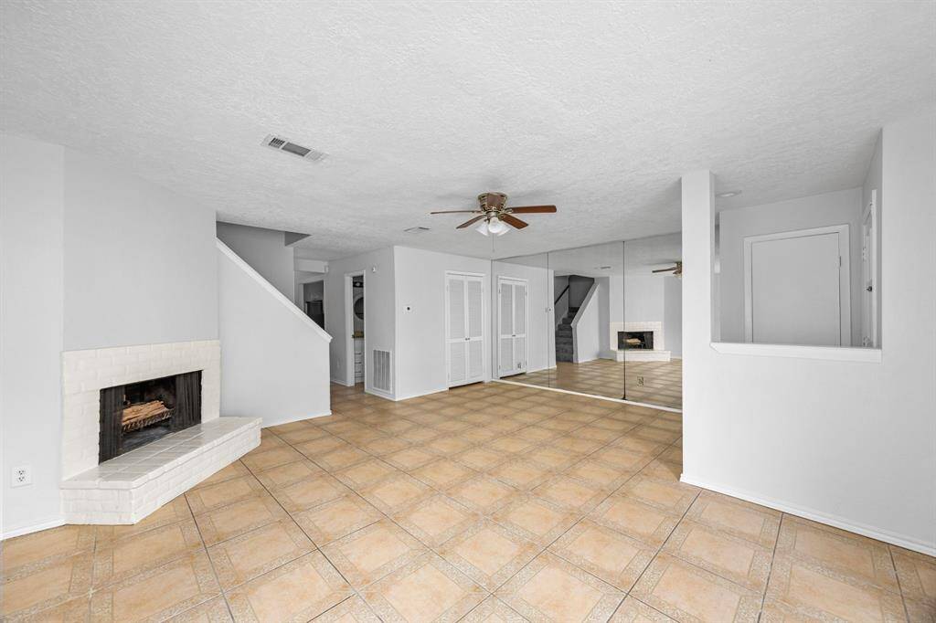 This spacious living area features a cozy white brick fireplace, tile flooring, and a ceiling fan. The mirrored wall enhances the room's brightness and sense of space, while an open stairway leads to the upper level.
