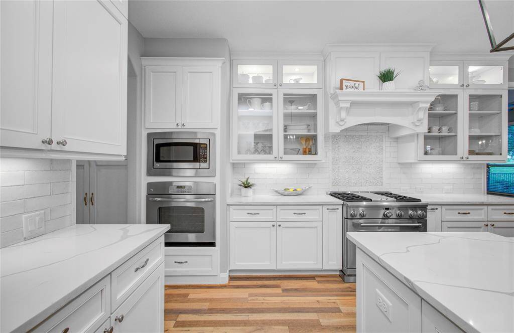 Bright, modern kitchen with custom cabinetry, stainless steel appliances, and a chef gas stovetop and double ovens. Glass-front cabinets, elegant backsplash, and wood flooring for a warm touch. Ideal for cooking and entertaining.